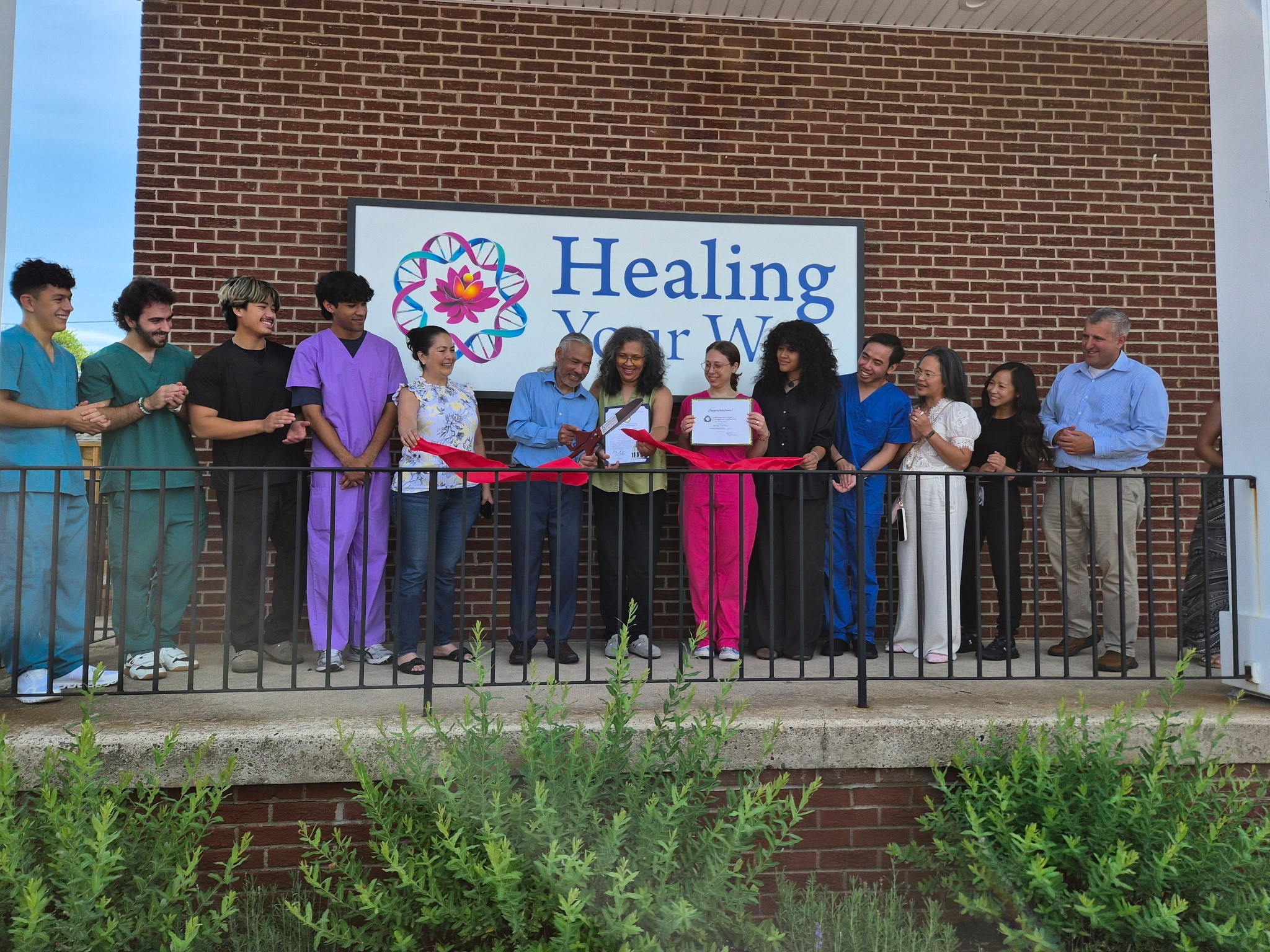 A group of people, including Sen. Farry, look on as a gentleman cuts a ribbon with giant scissors in front of Healing Your Way Building